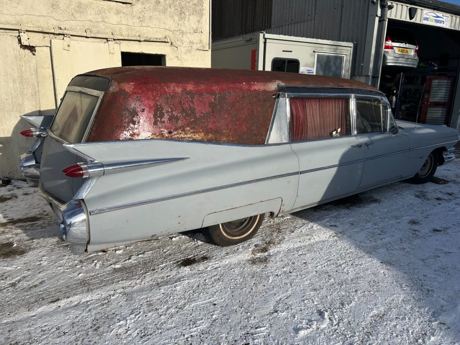 Ecto-1 on arrival in snowy Essex Classic hearse with a rusty red roof parked in a snowy lot.